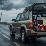 Un vehículo SUV estacionado en una carretera costera bajo una lluvia intensa, con el baúl abierto mostrando equipo de emergencia, suministros y el logo de LGV Auto Parts en la esquina superior.