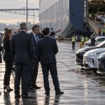 Ejecutivos observan vehículos de distintos fabricantes en un puerto de carga mientras autos importados descienden de un buque, en una escena que representa la competencia entre la industria automotriz estadounidense y los vehículos fabricados en China.